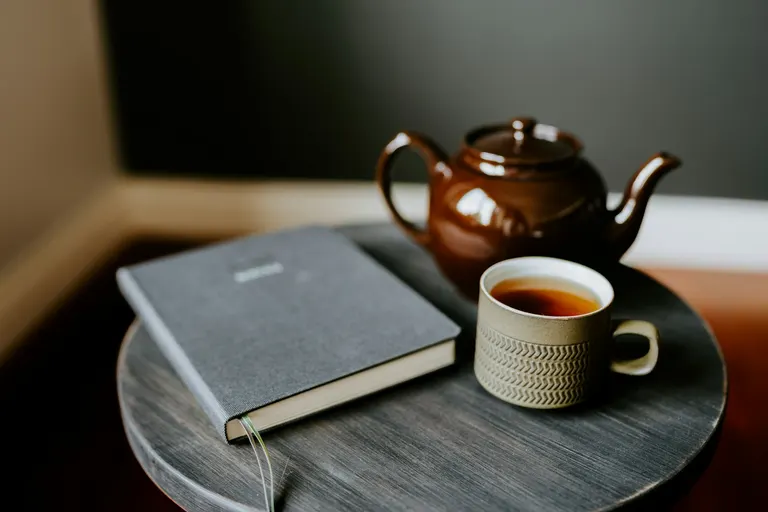 Een kopje thee, een theepot en een dichtgeslagen boek op een rond tafeltje in zacht licht.